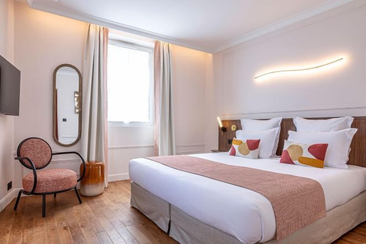 A hotel room with a bed, a chair, a mirror, and a window with curtains.