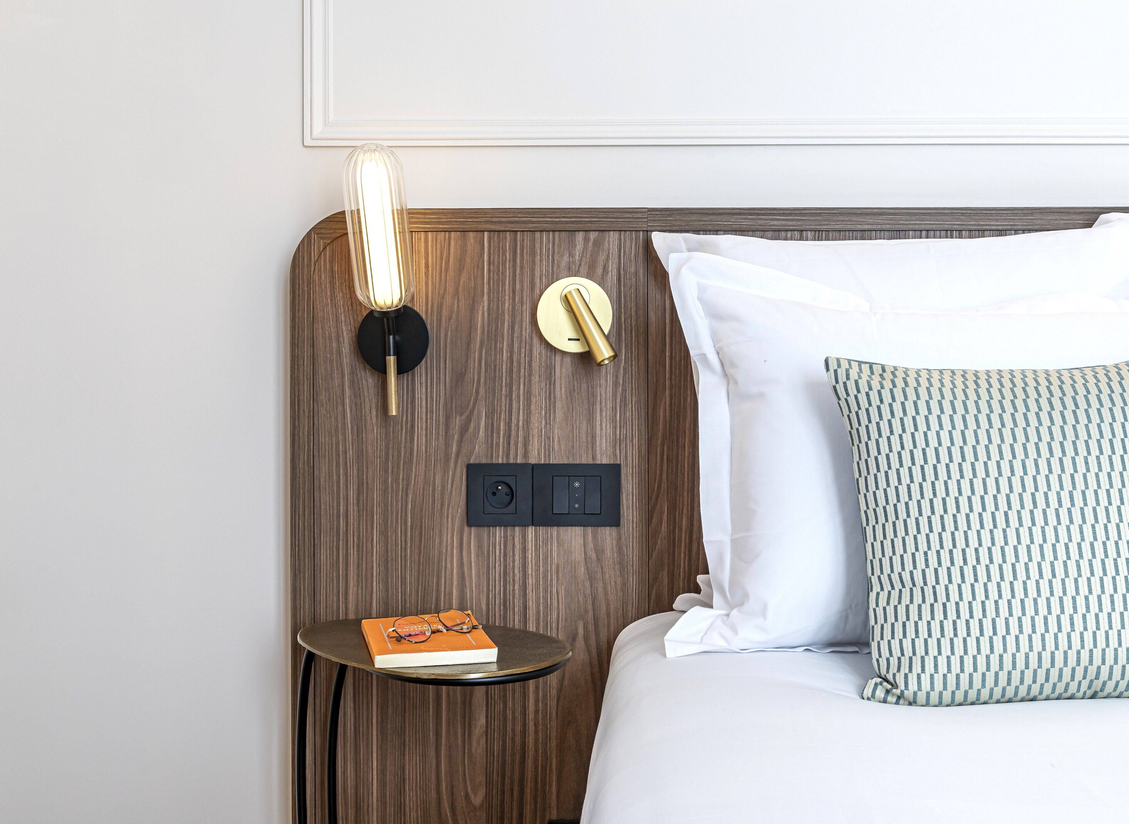A neatly made bed with white and patterned pillows, a bedside table with a book and glasses, a wall-mounted light fixture, and a thermostat.
