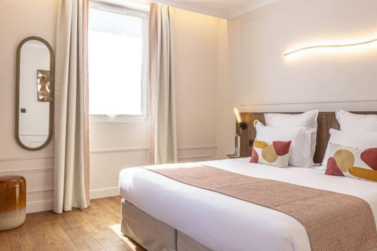 A hotel room with a large bed, a bedside table, a mirror, and a window with curtains.