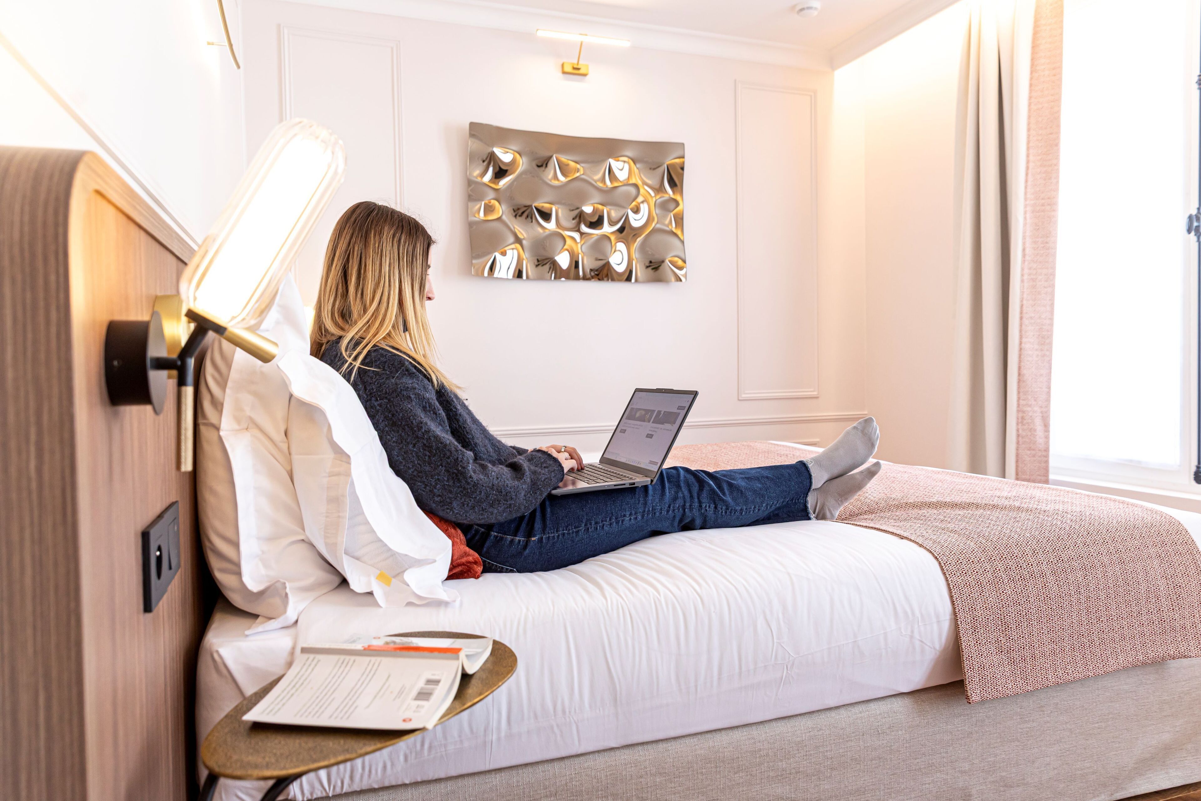 Een persoon zittend op een bed met een laptop, een nachtkastje met een lamp en een kunstwerk aan de muur.