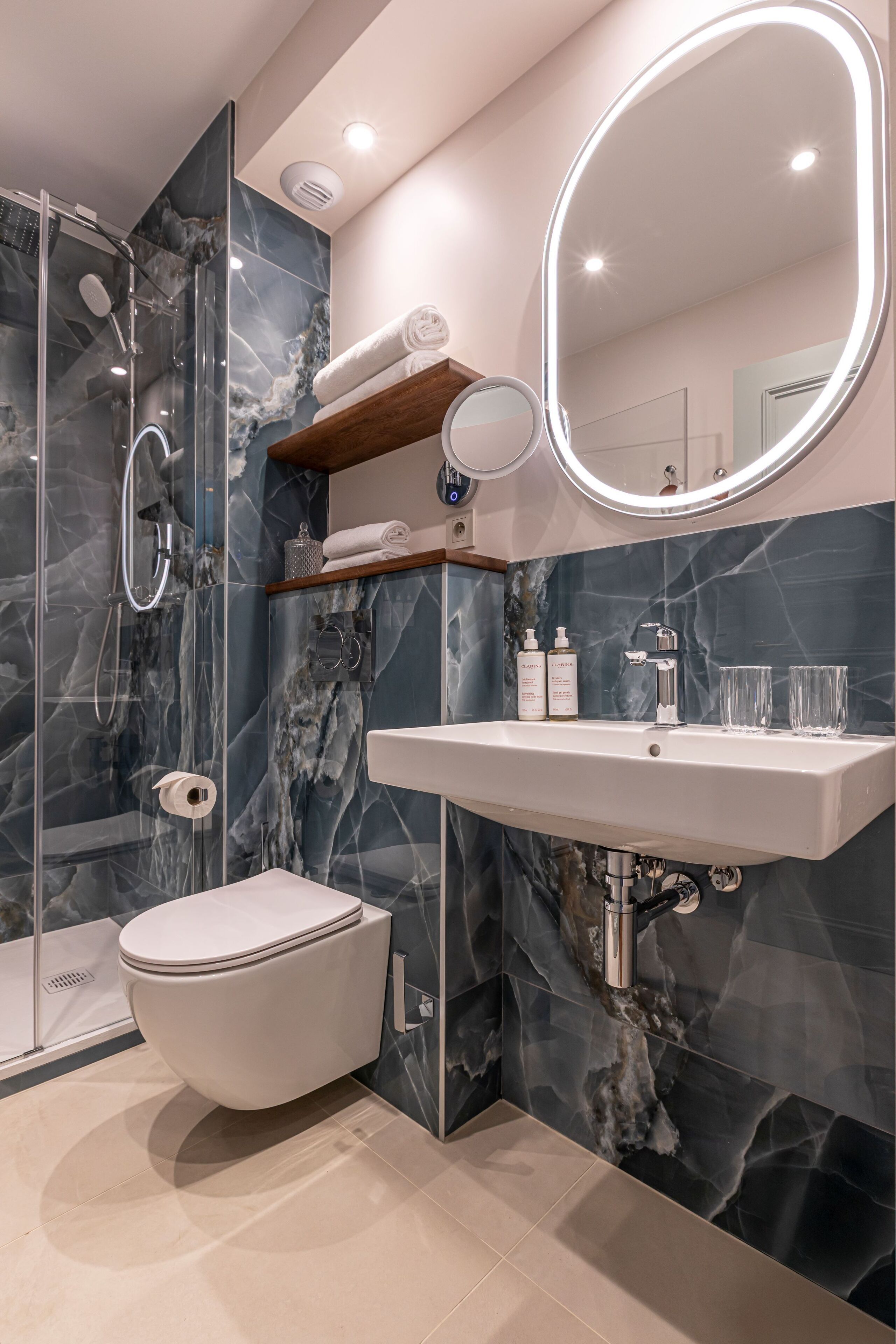 A modern bathroom with a marble shower, a toilet, a sink with a mirror, and a shelf with toiletries.