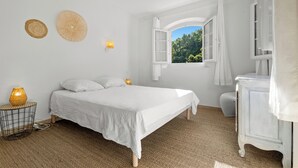 A bedroom with a bed, a bedside lamp, a small table, a window with curtains, and a view of greenery outside. - Vacation home, 250 m from the beach (Théoule-sur-Mer)