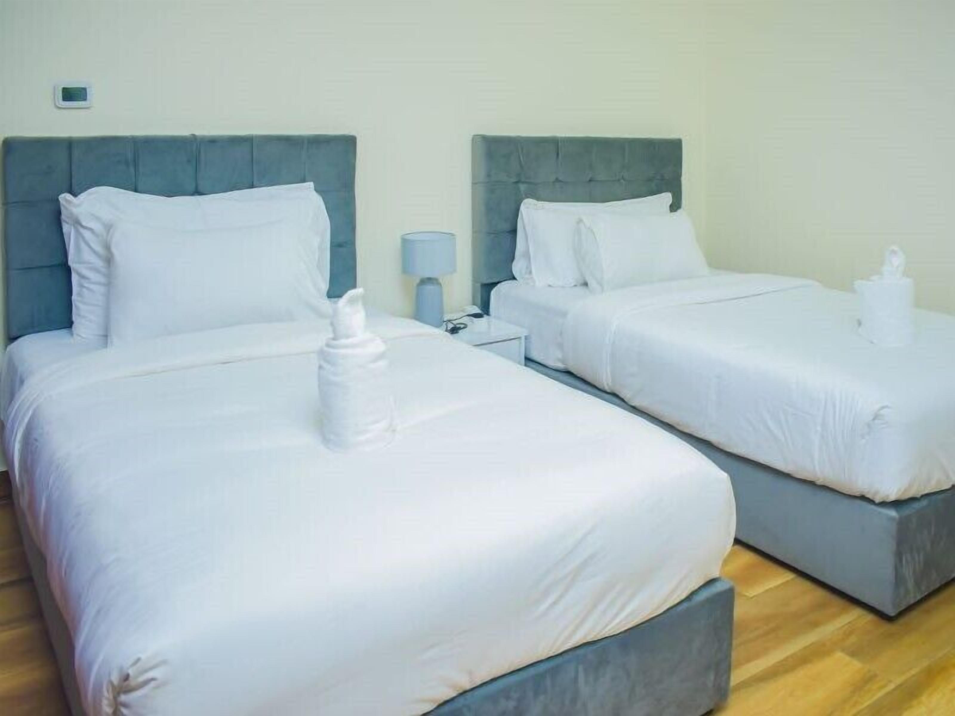 Habitación de hotel con dos camas, cada una con cabecero gris y ropa de cama blanca. En cada cama hay una mesita de noche con una lámpara y una toalla doblada.