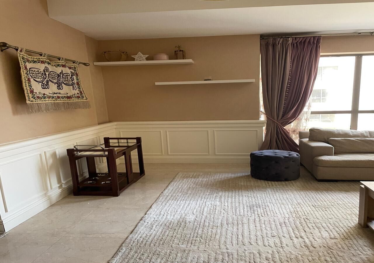 A living room with a beige sofa, a dark ottoman, a wooden chair, a rug, and a window with curtains.