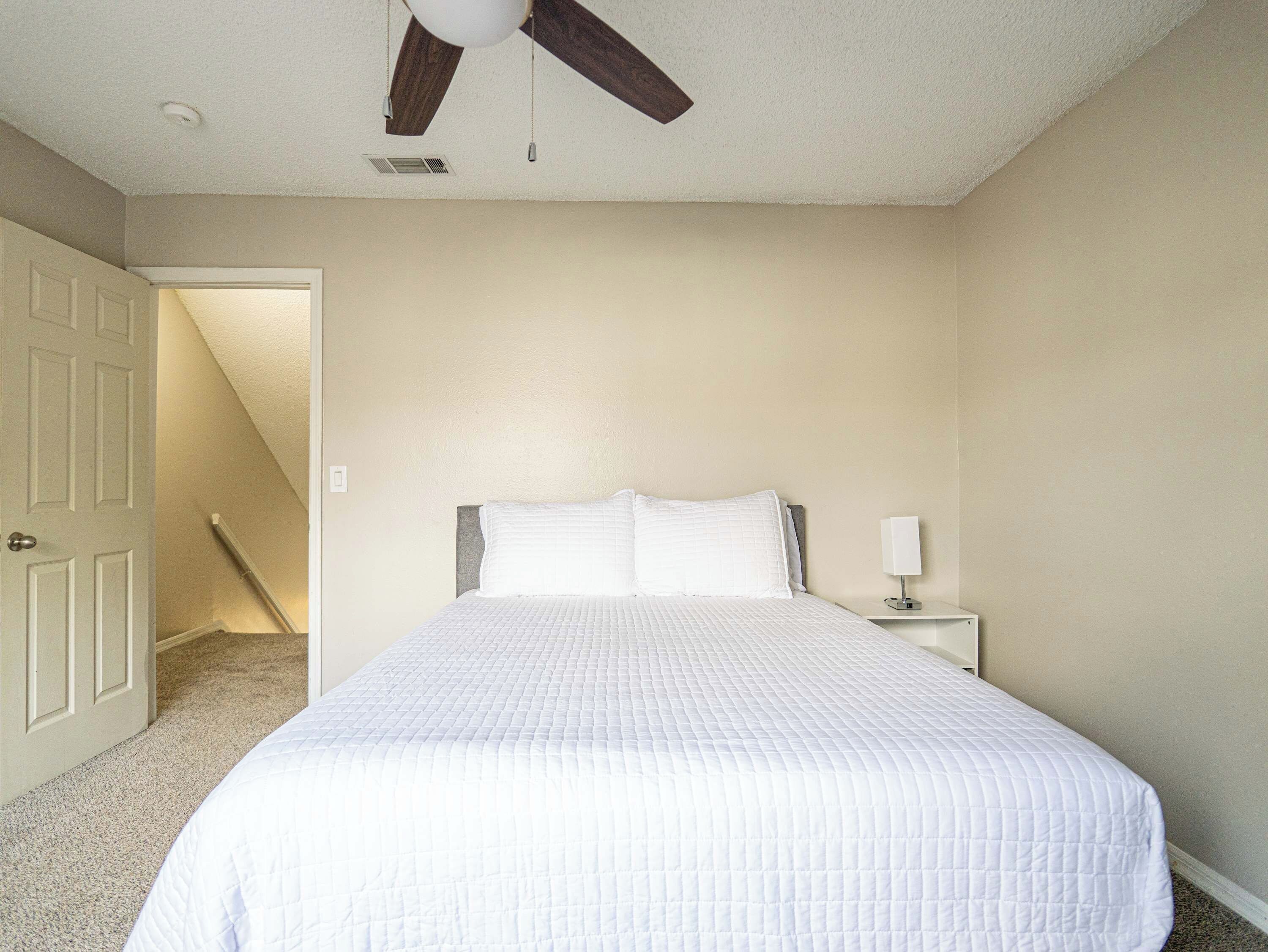 Une chambre à coucher comprenant un lit, une table de chevet, un ventilateur de plafond et une porte donnant sur une autre pièce.