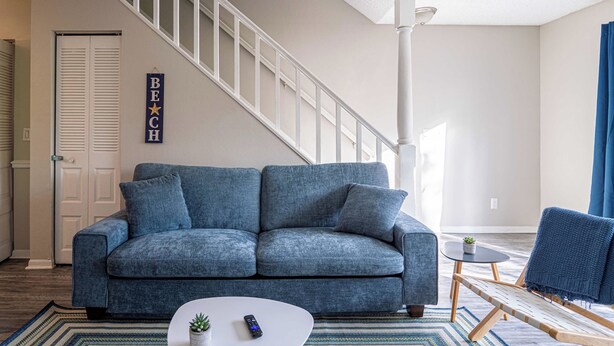 Ein Wohnzimmer mit einem blauen Sofa, einem weißen Couchtisch, einem kleinen Topfpflanzen, einer Fernbedienung und einem Deckenventilator.