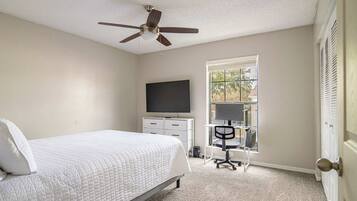 A bedroom with a bed, a desk with a chair, a ceiling fan, a TV, and a window.
