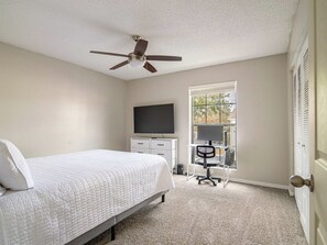 Une chambre à coucher avec un lit, un bureau et une chaise, un ventilateur de plafond, une télévision et une fenêtre.
