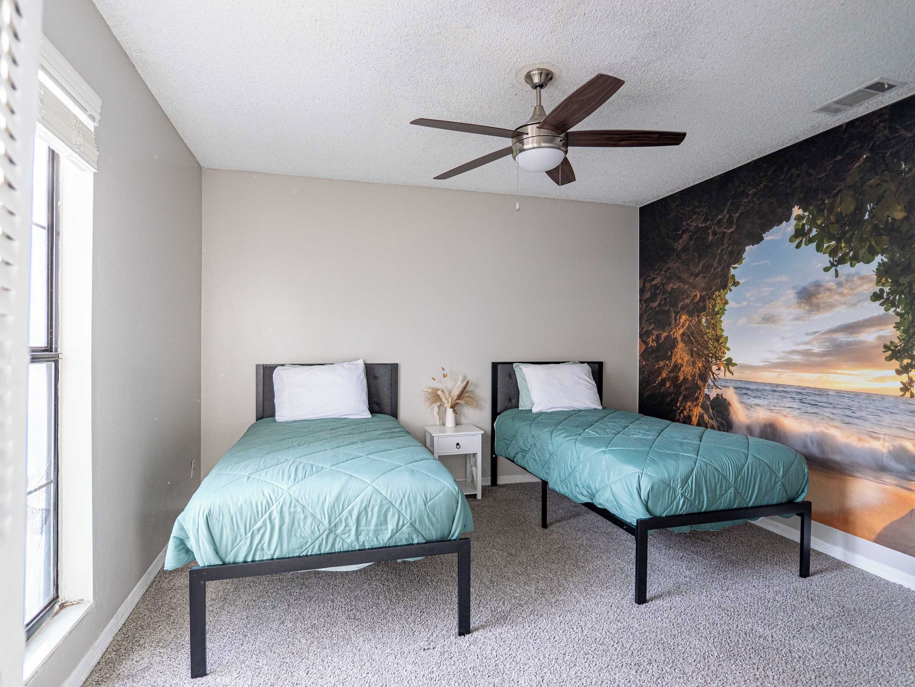 A bedroom with two beds, a ceiling fan, a nightstand, and a large mural of a sunset over the ocean.