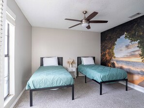Ein Schlafzimmer mit zwei Betten, einem Deckenventilator, einem Nachttisch und einem großen Wandbild, das einen Sonnenuntergang über dem Meer zeigt.