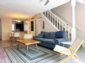Un salon avec un canapé, une table basse et un fauteuil. On y trouve un escalier avec une rampe et une porte sur laquelle est inscrit le mot « PLAGE ».