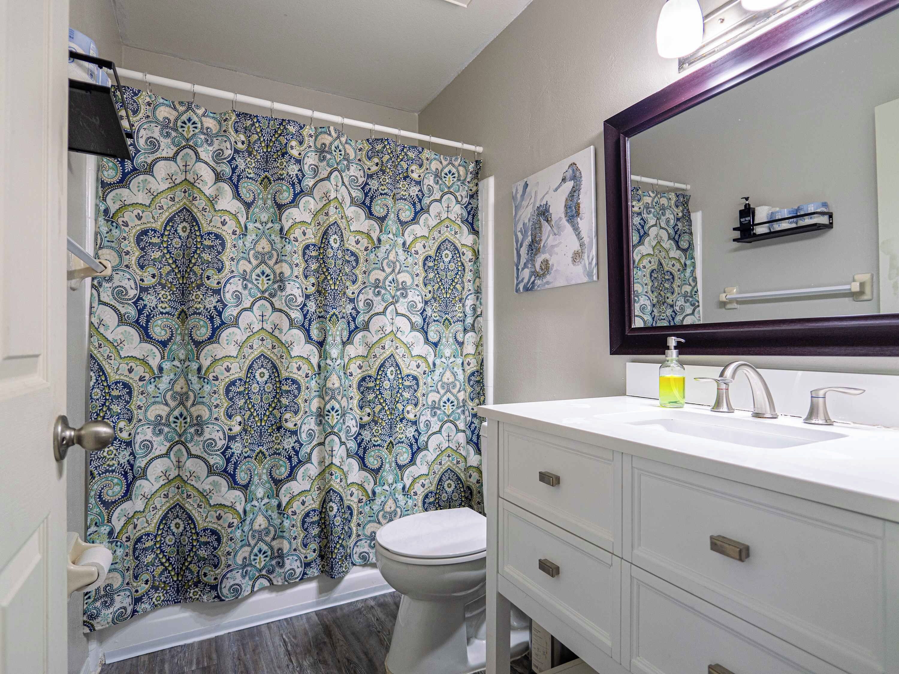 Une salle de bain avec un rideau de douche à motifs, des toilettes, un lavabo avec un miroir et un flacon de savon pour les mains.