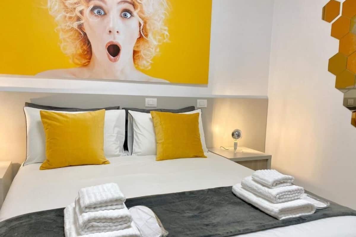 Et hotelværelse med en seng, to gule puder, en hvid pude, et natbord med en lampe samt en væg med et gult og orange design.