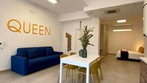 A modern interior with a white wall featuring the word "QUEEN" in gold letters, a blue sofa, a white dining table with yellow chairs, and a small plant. - BEEAPARTMENTS  (Bari)