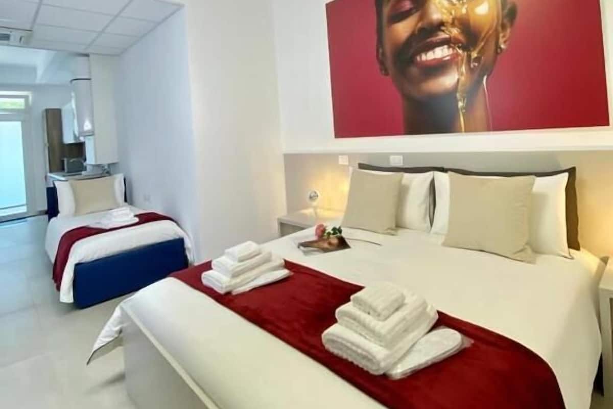 Et moderne hotelværelse med en stor seng, en mindre seng og et spiseområde, der ses igennem en åben dør.