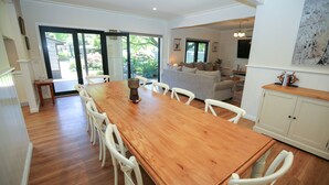A spacious dining area with a wooden table and white chairs, leading to a living room with a grey sofa and a television.