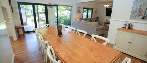 Un vaste espace dédié à la salle à manger, avec une table en bois et des chaises blanches, qui donne sur un salon doté d’un canapé gris et d’un téléviseur.