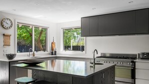 A modern kitchen with a large island, stainless steel appliances, and large windows offering views of greenery.