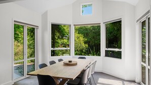 A modern dining room with a large table, multiple chairs, and large windows offering a view of greenery.