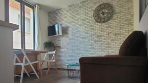 A living area with a brick wall, a clock, a TV, a sofa, and a dining table with chairs. - Cosy apartment, right in the center, 200m from the port  (La Ciotat)