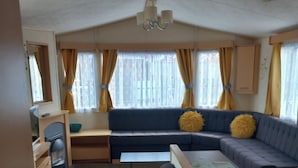 A compact living space with a sofa, a small table, a kitchenette, and a window with curtains. - Chapel St Leonards Happydays Beachfield (Skegness)