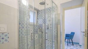 Une salle de bain avec une douche à parois de verre, des carreaux décorés, des toilettes et une chaise bleue dans la pièce adjacente.