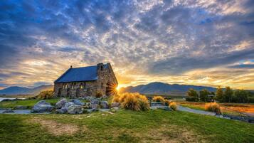 A stone building with a sloped roof, surrounded by grass and rocks, with mountains in the background.