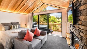 Ein modernes Schlafzimmer mit einem großen Bett, einem grauen Sofa mit roten Kissen, einem an einer Steinwand befestigten Fernseher und einer Glastür, die zu einem Außenbereich mit Whirlpool führt.