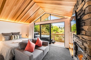 Et moderne soveværelse med en stor seng, en grå sofa med røde puder, et fjernsyn monteret på en stenvæg og en skydedør, der fører til et område med udendørs jacuzzi.