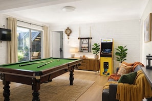 A room with a pool table, a Pac-Man arcade machine, a sofa, and a window with curtains. - Absolute Waterfront | Sleeps 10 | Family Retreat (Berkeley Vale)