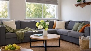 Een woonkamer met een grijze bank, een ronde salontafel, een vaas met bloemen en een schaal fruit.