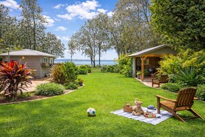 A well-maintained lawn with a picnic setup, a wooden chair, and a small building with a covered porch. - Absolute Waterfront | Sleeps 10 | Family Retreat (Berkeley Vale)