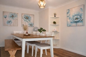 A dining area with a wooden table, white chairs, a bench, and wall-mounted shelves with decorative items. - Absolute Waterfront | Sleeps 10 | Family Retreat (Berkeley Vale)