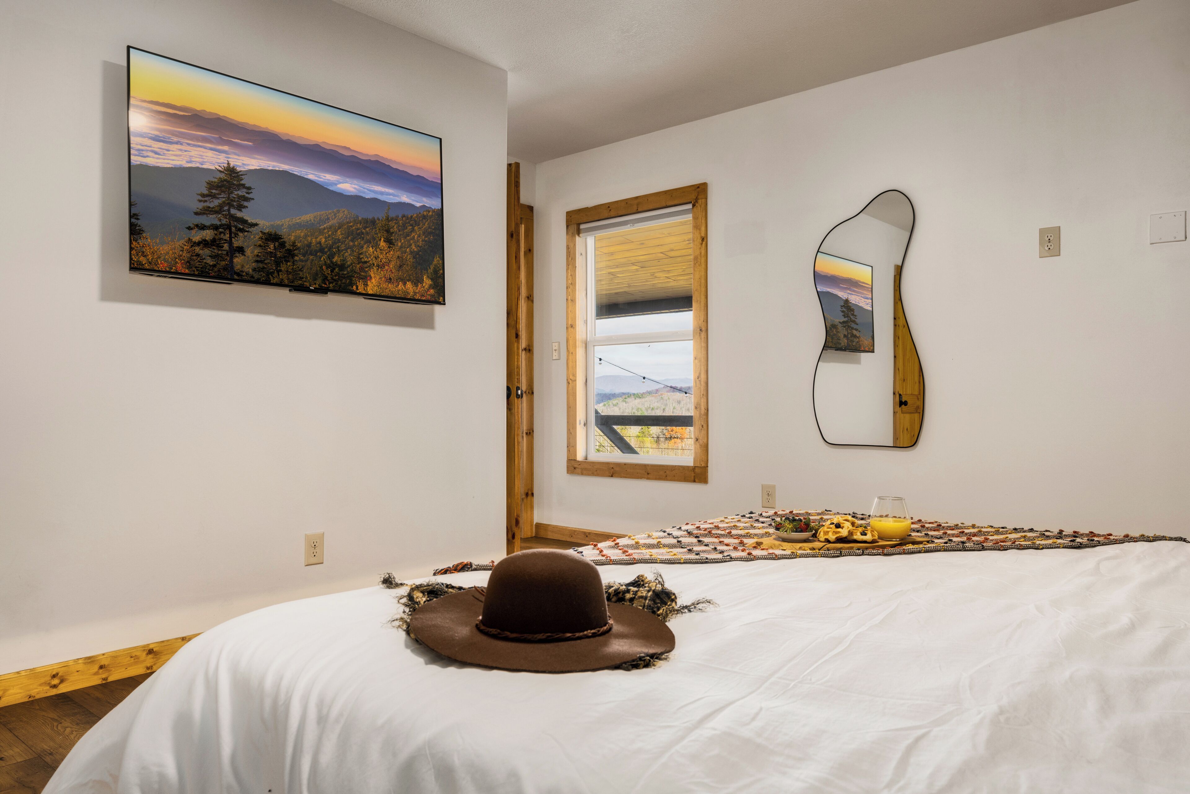 Ein Schlafzimmer mit Bett, Hut auf dem Bett, Landschaftsbild, Fenster und Spiegel an der Wand.