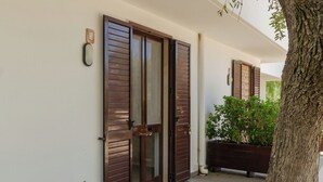 Un edificio de fachada blanca, una puerta de madera con paneles de vidrio y un árbol en primer plano.