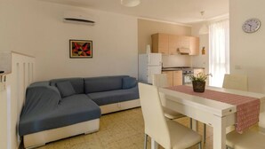 Una sala de estar moderna con un sofá seccional gris, una mesa de comedor blanca con camino de mesa rojo, y una zona de cocina con armarios de madera y un refrigerador blanco.