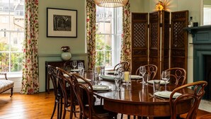 A dining room with a round wooden table set for six, flanked by tall wooden chairs. A large, woven pendant light hangs above the table, and a framed artwork is displayed on the wall. - Grand Georgian Townhouse With Garden (Bath)