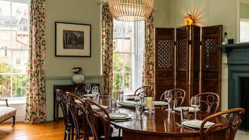 A dining room with a round wooden table set for six, flanked by tall wooden chairs. A large, woven pendant light hangs above the table, and a framed artwork is displayed on the wall.