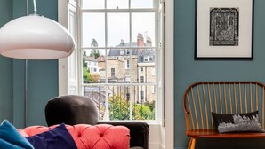 A room with a large window, a black sofa, a wooden chair, and a framed artwork on the wall. - Grand Georgian Townhouse With Garden (Bath)