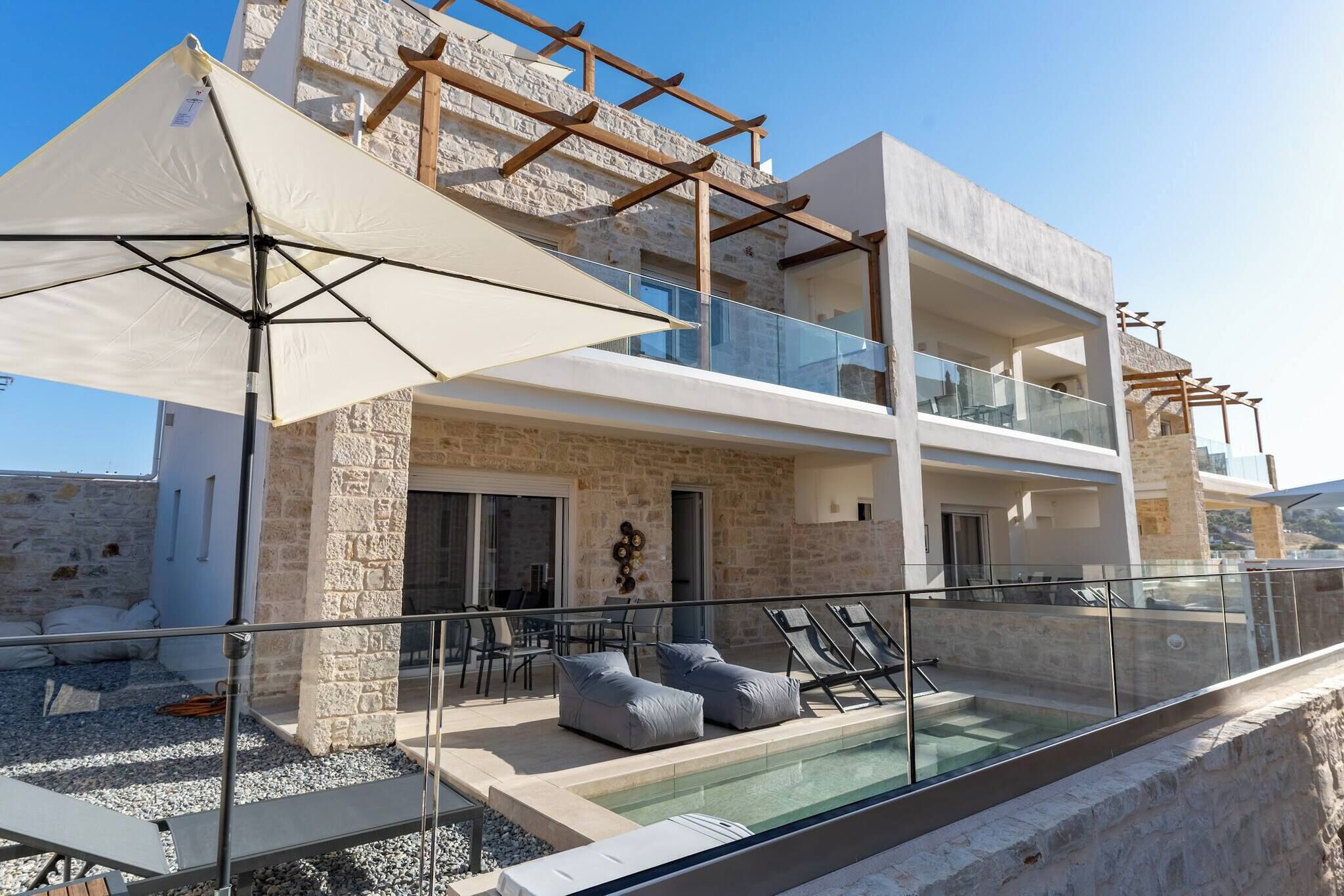 A modern two-story building with a stone facade, a glass balcony, and a swimming pool area with lounge chairs.