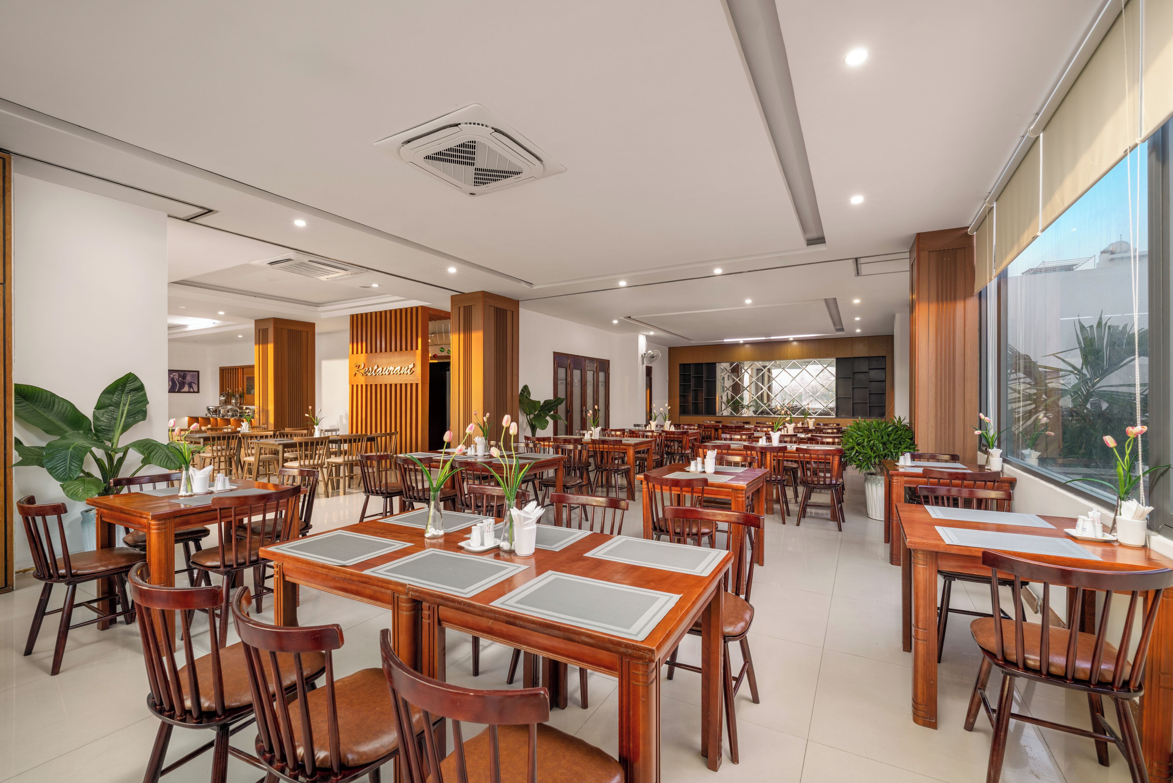 A spacious dining area with multiple wooden tables and chairs, large windows, and indoor plants.
