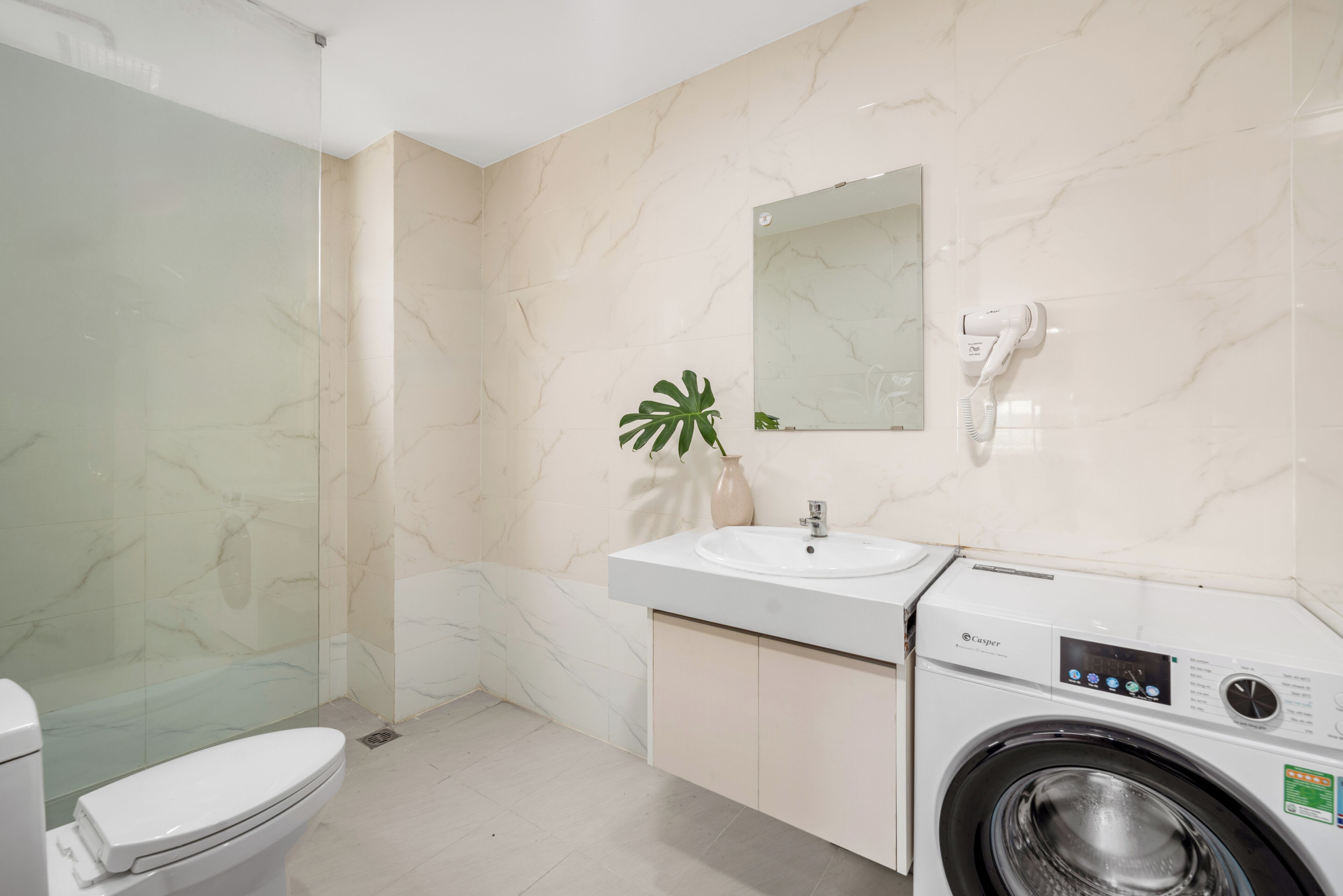 A modern bathroom with a glass shower enclosure, a white sink, a mirror, and a front-loading washing machine.