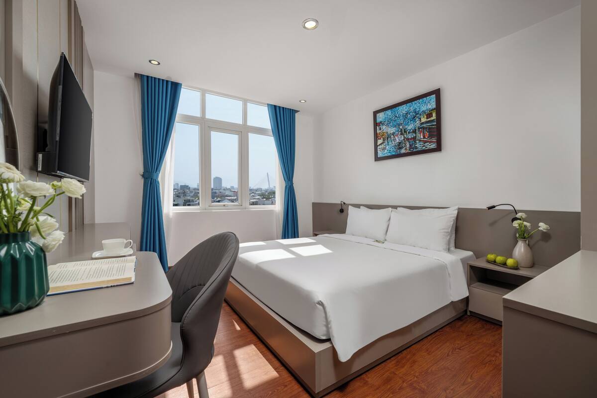 Una habitación de hotel moderna con cama, escritorio, silla y un amplio ventanal con cortinas.
