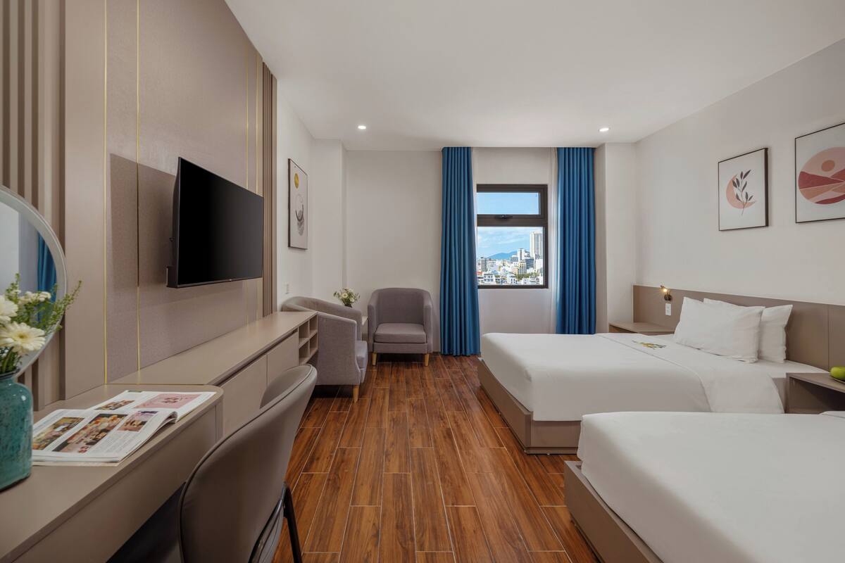 Habitación de hotel con televisión, escritorio con un florero, una silla, cama y ventana con cortinas.