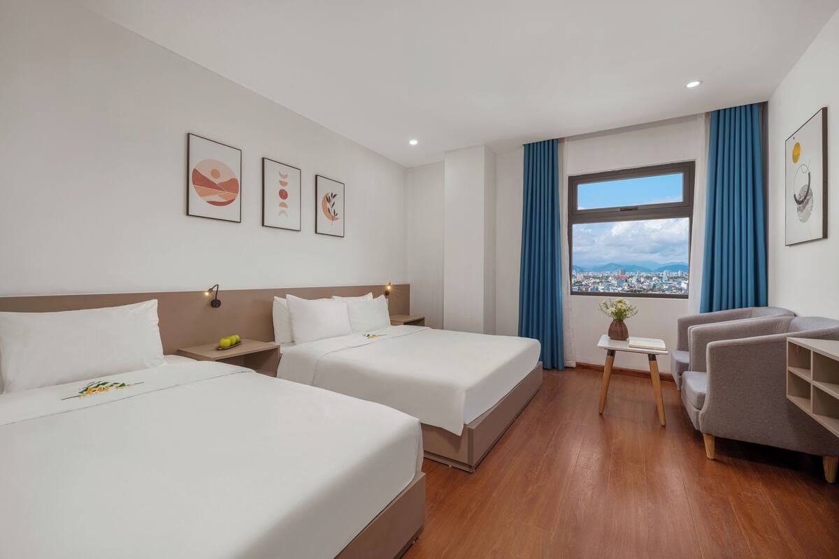 Habitación de hotel con dos camas, una mesita con un florero, un sofá y un ventanal con vistas a la ciudad.