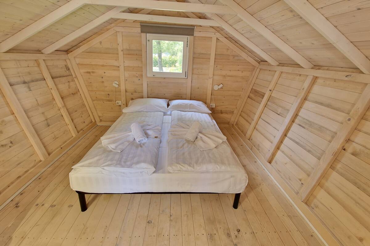 Een gezellig slaapkamer onder het dak, met een houten schuine plafond, een enkel bed met witte lakens en een klein raam dat natuurlijk licht biedt.