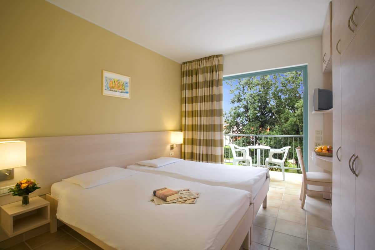 Ein Hotelzimmer mit zwei Betten, einem Balkon mit Tisch und Stühlen sowie einem Fernseher auf einem Schrank.
