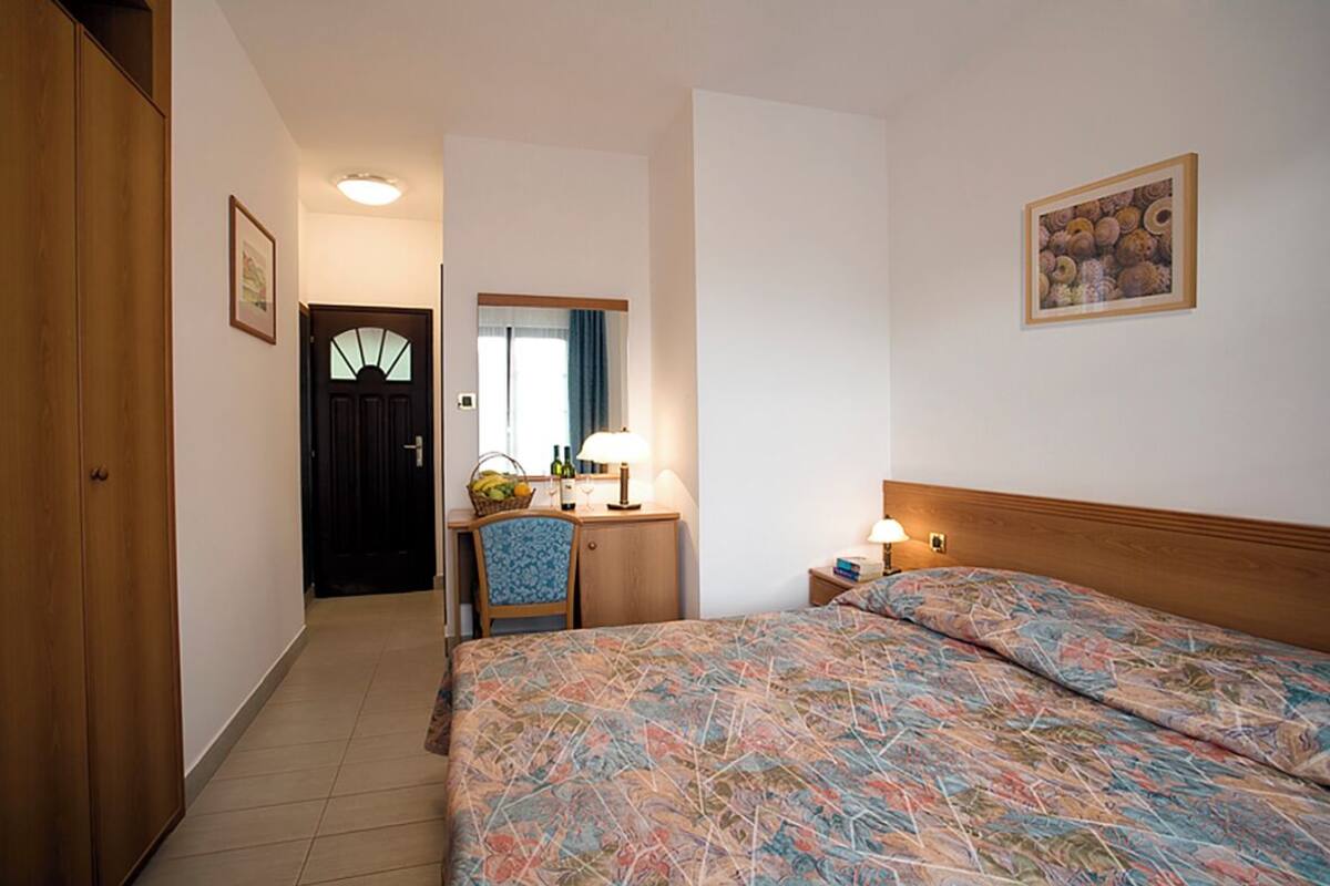 Een hotelkamer met een bed, een bureau met een lamp, een stoel en een deur naar een andere kamer.