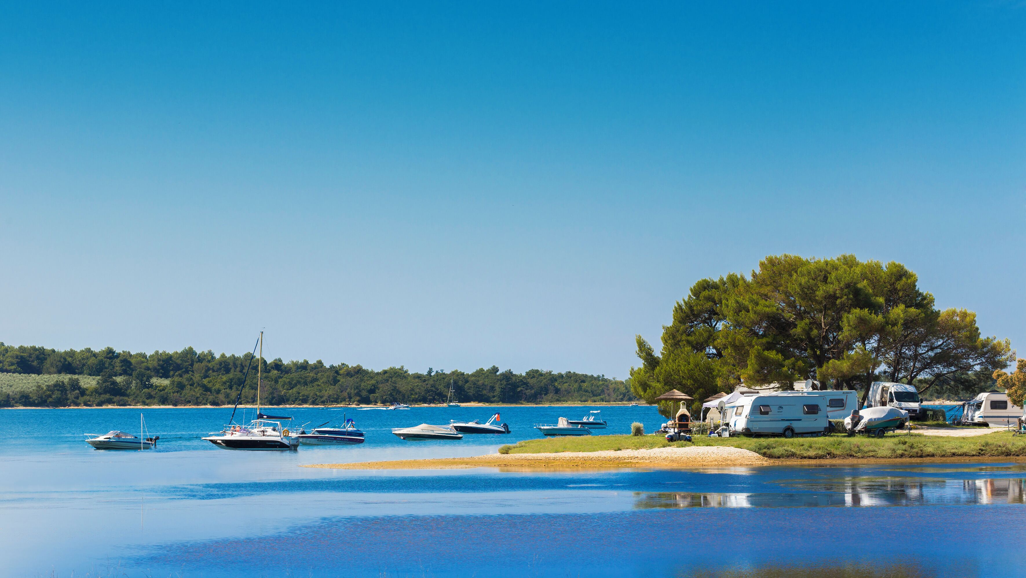 Um parque de campismo à beira do lago, com várias embarcações, veículos recreativos e uma grande árvore a dar sombra.