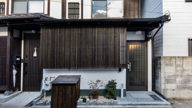 A traditional Japanese building with a wooden entrance, a sliding door, and a small garden area with a wooden bench and potted plants. - Shiki Homes MEIGETSU (Kyoto)